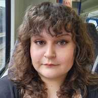 A selfie of Kerstin. Kerstin has short, brown curly hair and brown eyes. She is wearing a black cardigan and a black and white top. She is looking at the camera and appears to be sitting on a train.