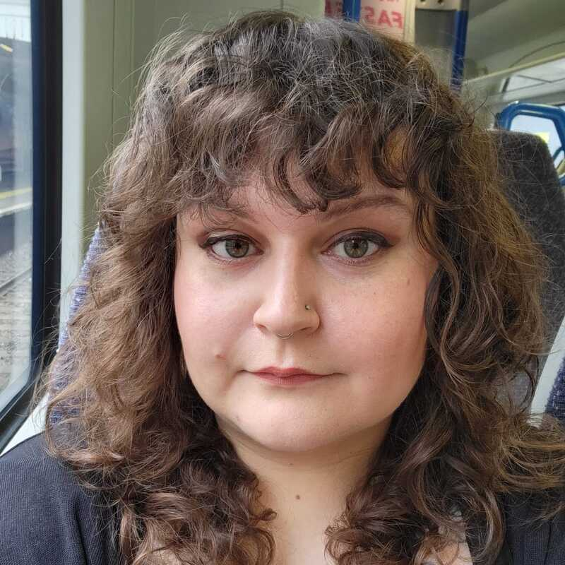 A selfie of Kerstin. Kerstin has short, brown curly hair and brown eyes. She is wearing a black cardigan and a black and white top. She is looking at the camera and appears to be sitting on a train.