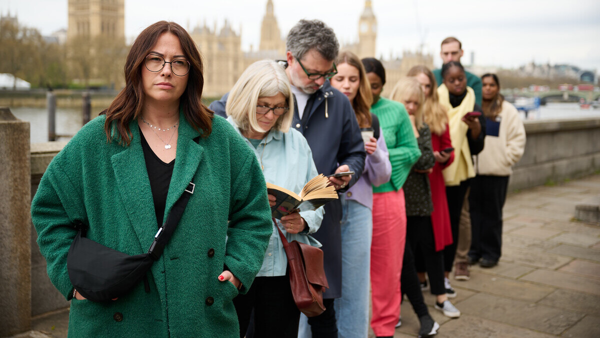 A group of people are standing in a queue on a bridge near the Parliament building in London. The person in front of the queue has short brown hair and is wearing a green jacket. Behind them are people looking at their phones, books and other objects.