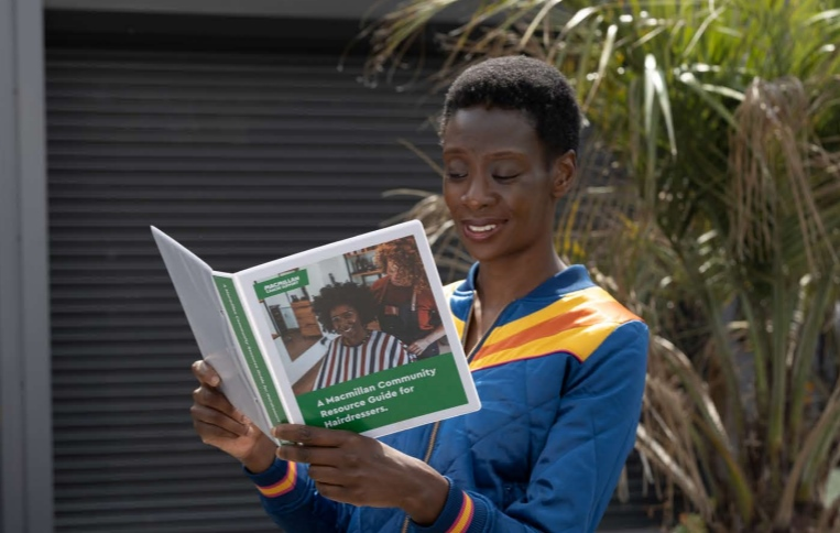 A black women with short hair is smiling as she reads a booklet called A Macmillan Community Resource Guide for Hairdressers