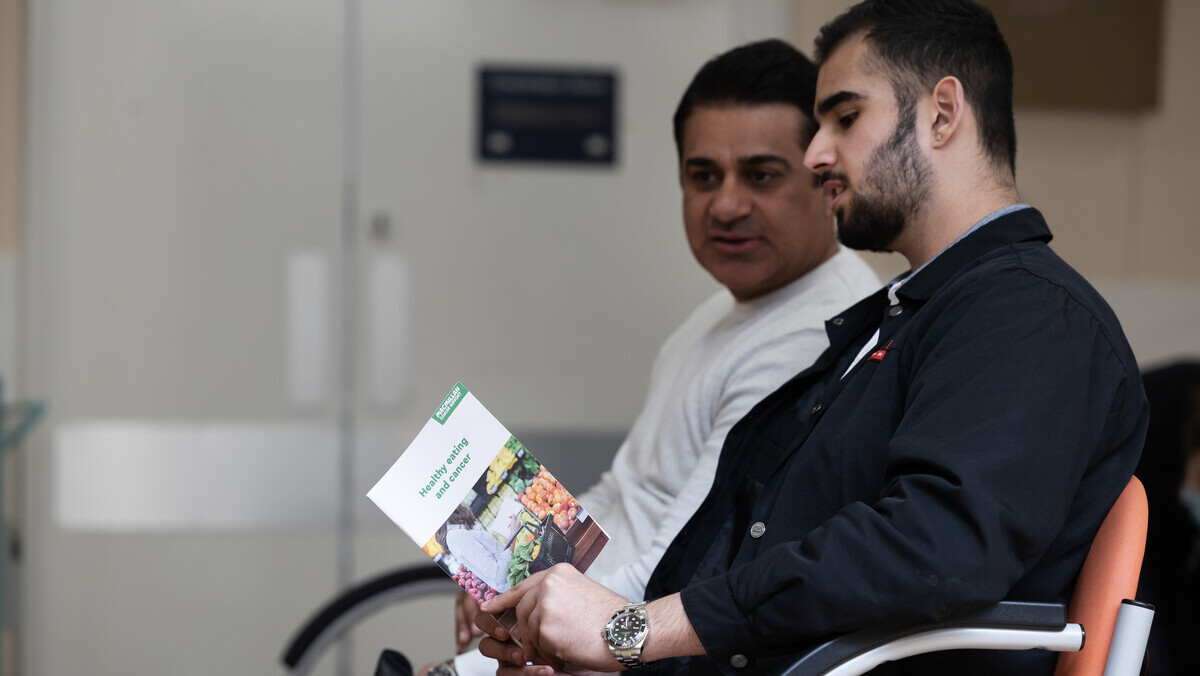 Jai and Mandeep are sitting in chairs in a waiting area. Jai is wearing a long, dark coat and is holding open a booklet. Next to him is Mandeep who is wearing a long sleeved white top. Both appear to be talking to each other. 