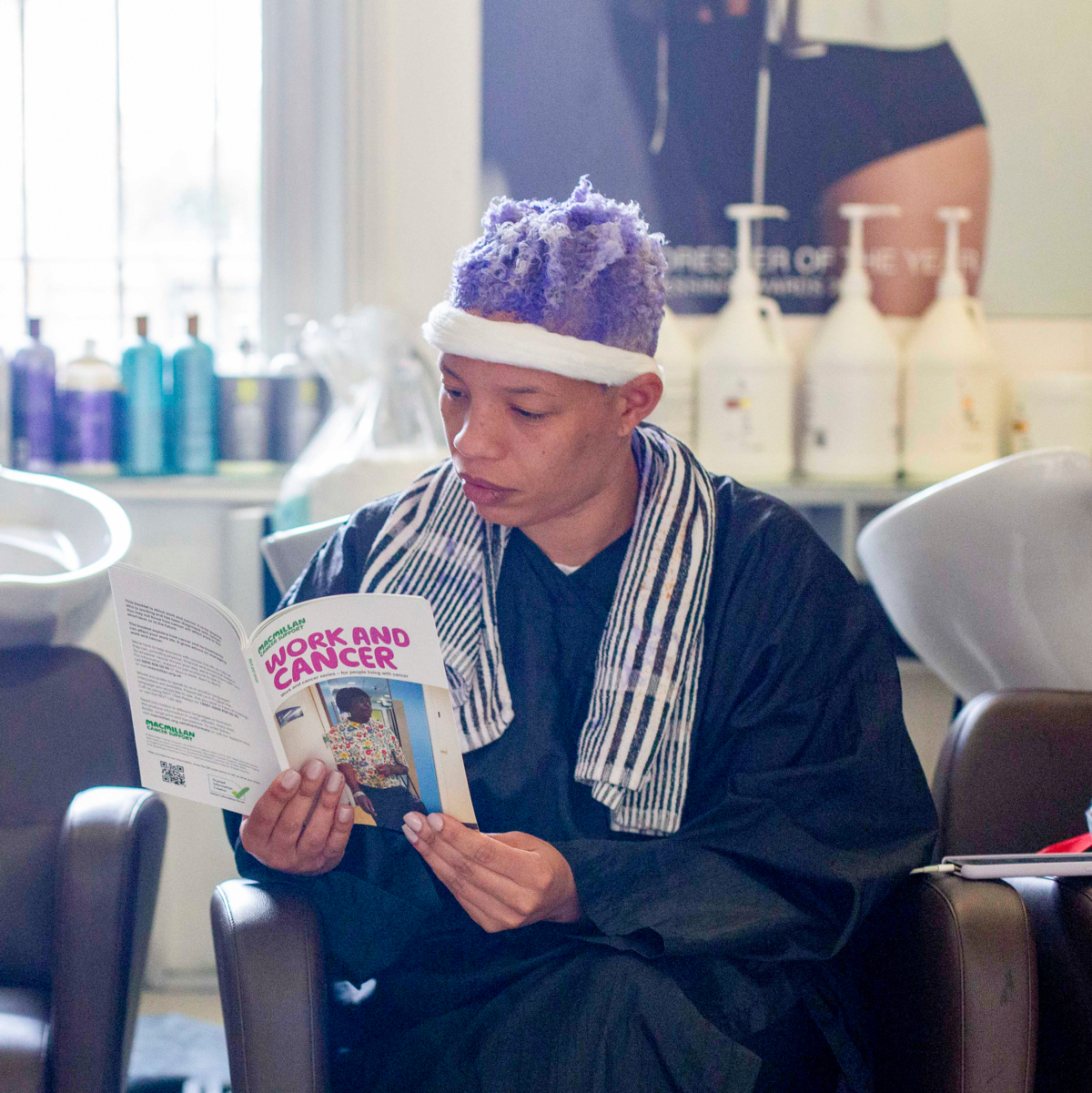 A person is sitting by the sink in a hairdressers reading a booklet about work and cancer from Macmillan