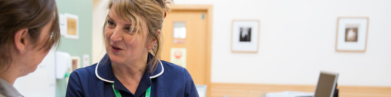 Shez, wearing her dark blue nurse's uniform and a green Macmillan lanyard, is talking to another nurse. They are in discussion.