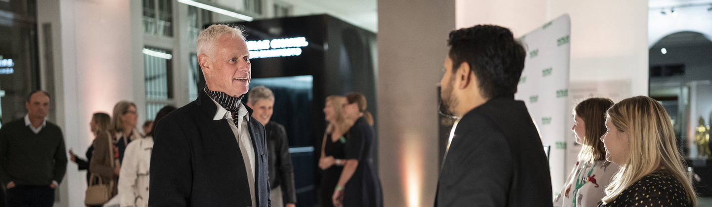 A man is speaking to a person standing behind a table. The man is wearing a suit and a blue and white striped ascot. On the table are name tags. 