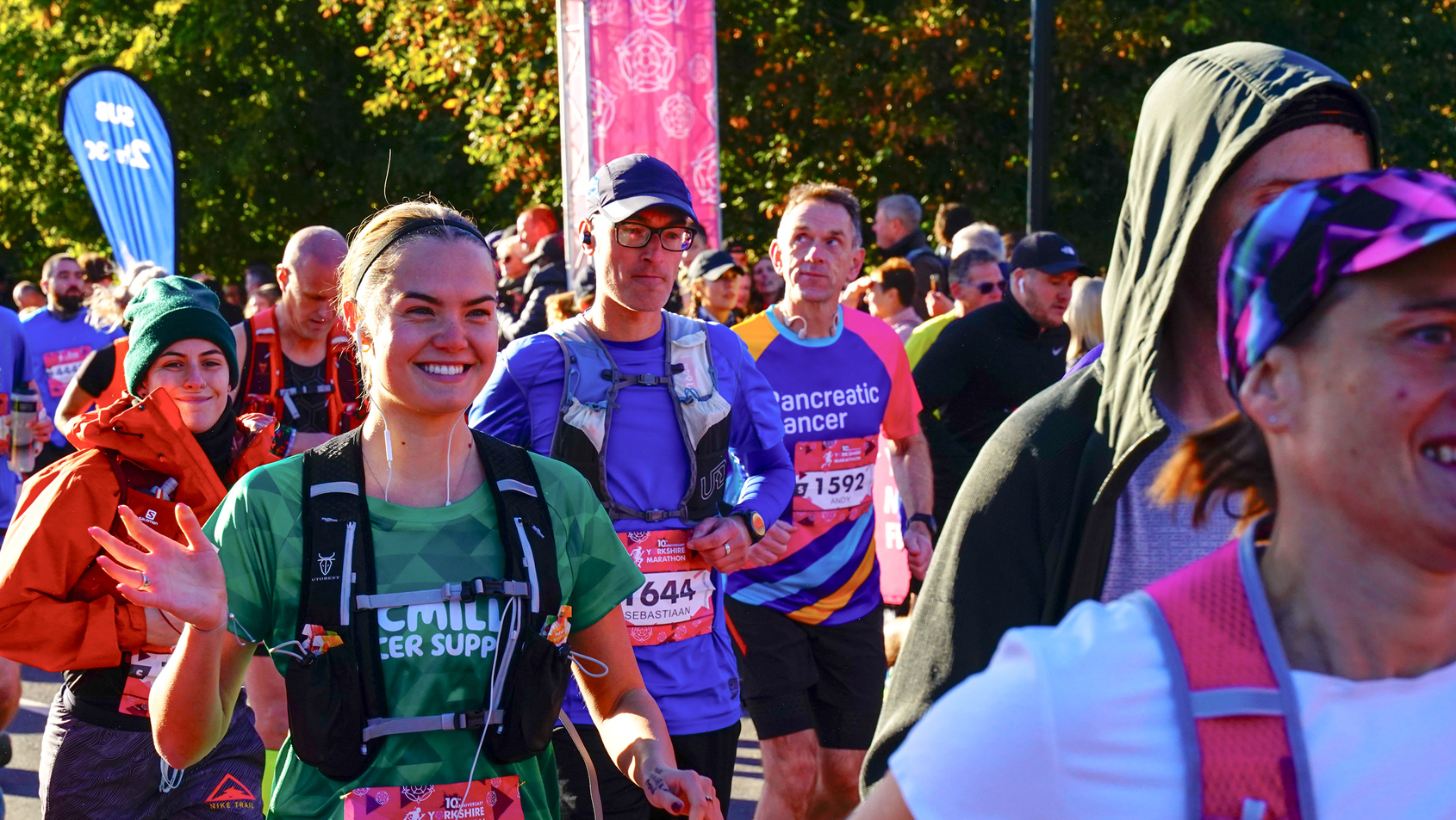 A person running at an event. The person is wearing a green Macmillan running top and a black running vest. Their brown hair is tied back. Around them are other runners.