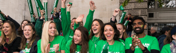 A group of volunteers cheering at an event. They are standing behind a green barrier. They are wearing green Macmillan tops and some are holding Macmillan branded cheering balloons. 