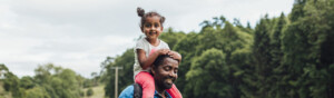 A father and daughter are walking in a field. The daughter is on top of the father's shoulders. Behind them is green grass and sheep.