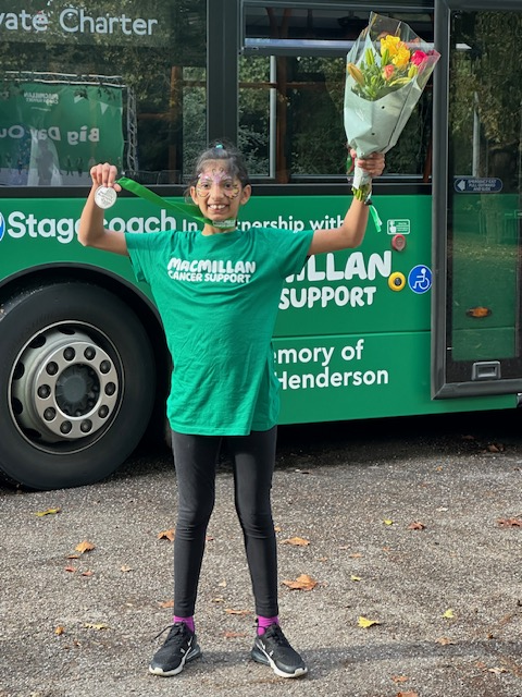 Young girl wearing a Macmillan t-shirt and posing with a medal and a bunch of flowers in front of a Macmillan double decker bus