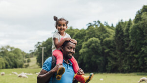A father and daughter are walking in a field. The daughter is on top of the father's shoulders. Behind them is green grass and sheep.
