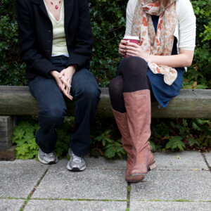 Two women sitting on a bench talking. One is wearing brown boots and a floral scarf the other is wearing a black cardigan, jeans and trainers.