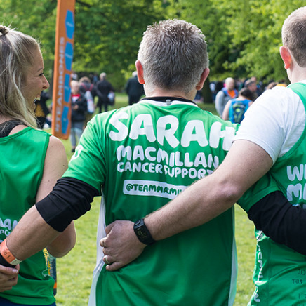 Three people photographed from behind with their arms around each other wearing green Macmillan running tops