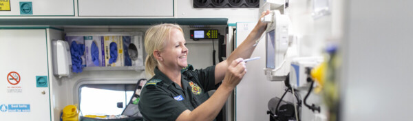 A paramedic is checking a screen in an ambulance. The paramedic has their blonde hair pulled back and is wearing a green paramedic outfit. Around them is medical equipment. 