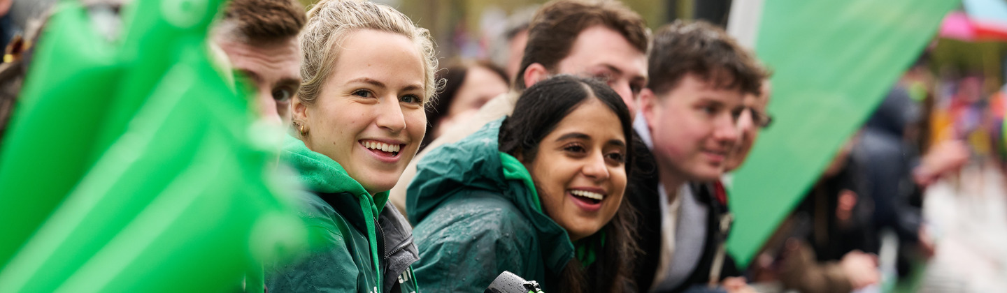 A group of volunteers are cheering at the London Marathon. The volunteers are at a cheer point. Some are holding long Macmillan branded cheer balloons. One volunteer has a green megaphone. 