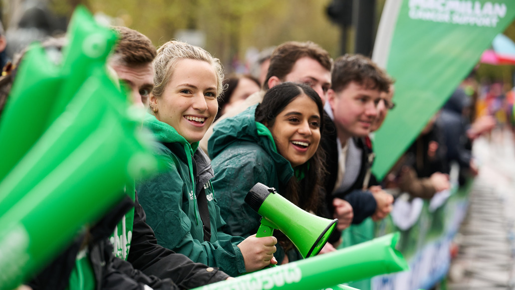 A group of volunteers are cheering at the London Marathon. The volunteers are at a cheer point. Some are holding long Macmillan branded cheer balloons. One volunteer has a green megaphone. 
