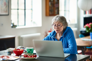 An older woman sits at her kitchen table working on a laptop
