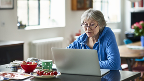 An older woman sits at her kitchen table working on a laptop