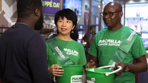Two Macmillan volunteers are talking to a person in a Costa. Both volunteers are wearing green Macmillan logo t-shirts and sashes. They are both holding green collection buckets. The person they are speaking to is wearing a dark blue top. 