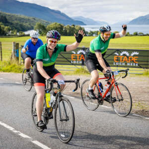 Two cyclists riding in the Etape Loch Ness event, smiling and waving at the camera. 