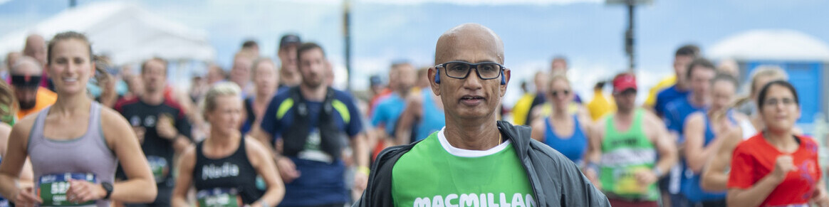 The runner is wearing a green Macmillan running top. They are also wearing a grey jacket and gloves, and blue shorts. Behind them are others runners. 