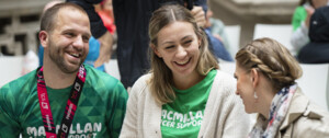 Three people are sitting down and talking. The person on the left is wearing a Macmillan running top and a metal from the London Marathon. The person in the middle is wearing a Macmillan top and the person on the right is wearing a trench coat.