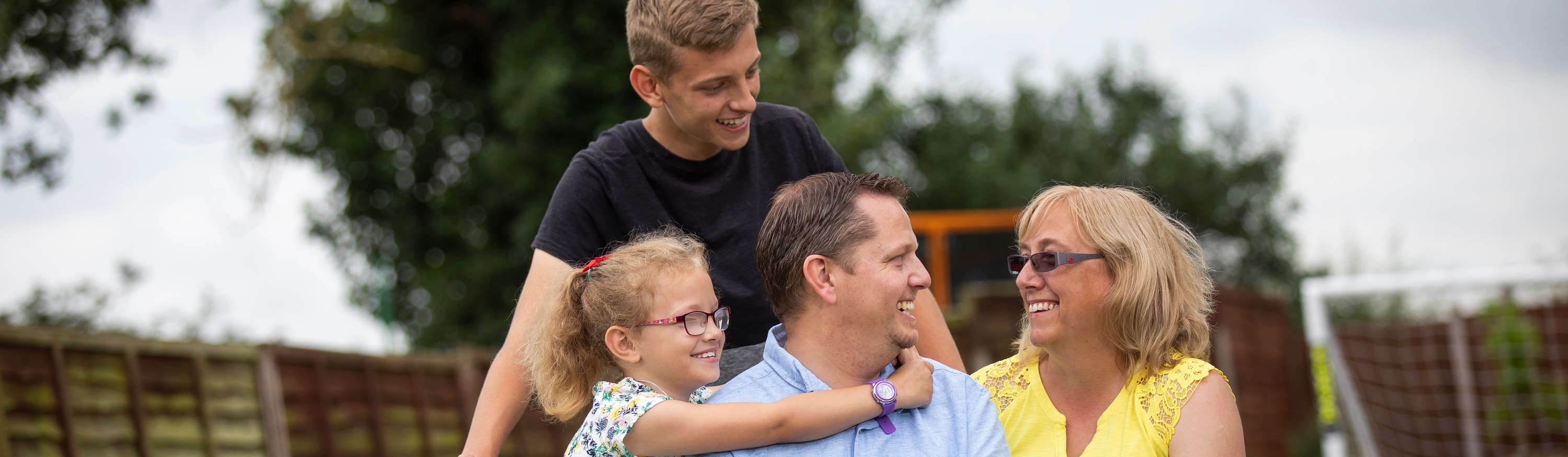 A man and woman sitting in the garden with their two children, a boy standing behind them and a girl with her arms around the man