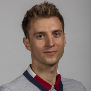Man smiling at camera, he is wearing a grey top with a red, blue and white stripe collar.