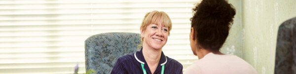 Nurse in blue uniform and green lanyard talking to woman. They're sitting on chairs facing on another.   