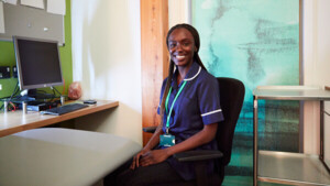 A nurse is sat in an office chair and facing towards the camera smiling. She has long braids in her hair, is wearing a blue nurse's uniform, and has on a green lanyard. Next to her is a desk with a computer. 