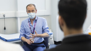 Dr Grumett is sitting down in a chat. He is wearing a face mask, a long blue shirt, blue trousers and a purple lanyard. He appears to be speaking to a patient.