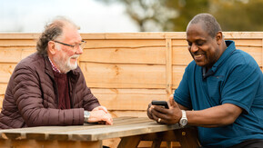 Two men sitting across from each other on a picnic bench talking.