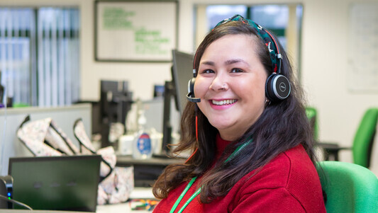 Jenny is a Macmillan Support Line Welfare Rights Advisor. She is sitting at a desk and smiling at the camera. She is wearing a red top and blue jeans. On her head is a headset and she appears to be working at a computer. 