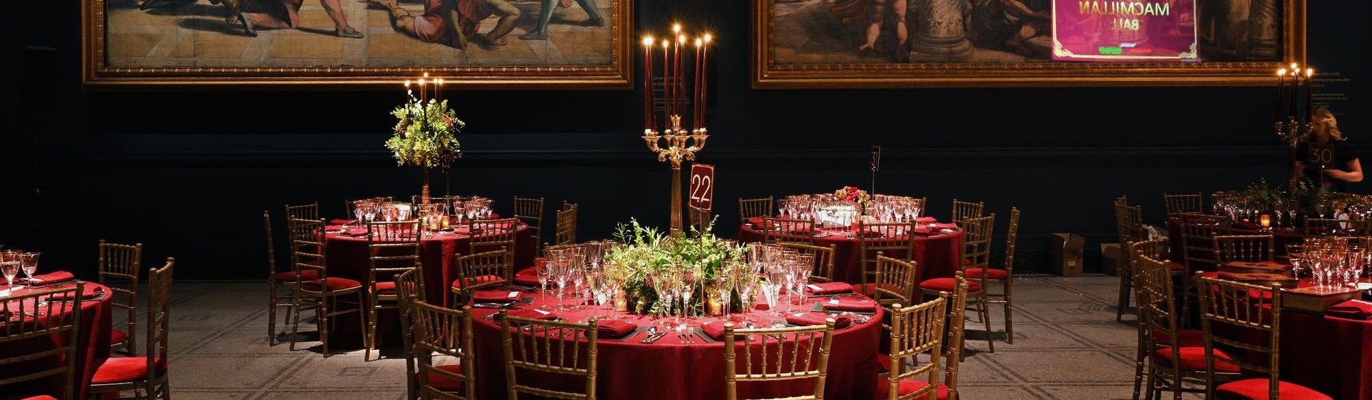 Several tables and chairs have been set up in a art gallery for an event. The tables are decorated with red table cloths, a candelabra, and place settings for dinner. On the walls hangs different pieces of artwork. 