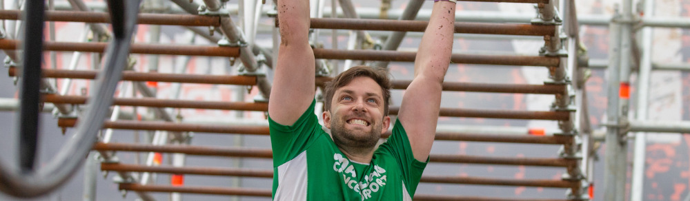 A man with short brown hair is moving across monkey bars. He is wearing a green Macmillan top. Behind him are other competitors waiting to use the monkey bars. It appears these have been set up outside.
