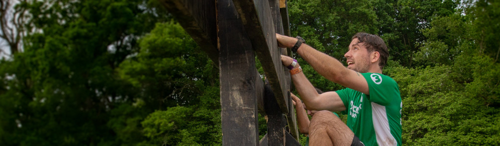 A man is climbing up a wooden structure at a Spartan race. He has short brown hair and is wearing a green Macmillan top and dark shorts. Behind him are tress and grass. 