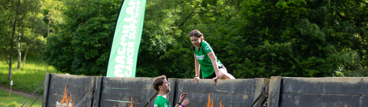 A woman with long brown hair that has been braided is climbing over a wooden wall at an obstacle event. She is wearing a green Macmillan top and appears to be outside.