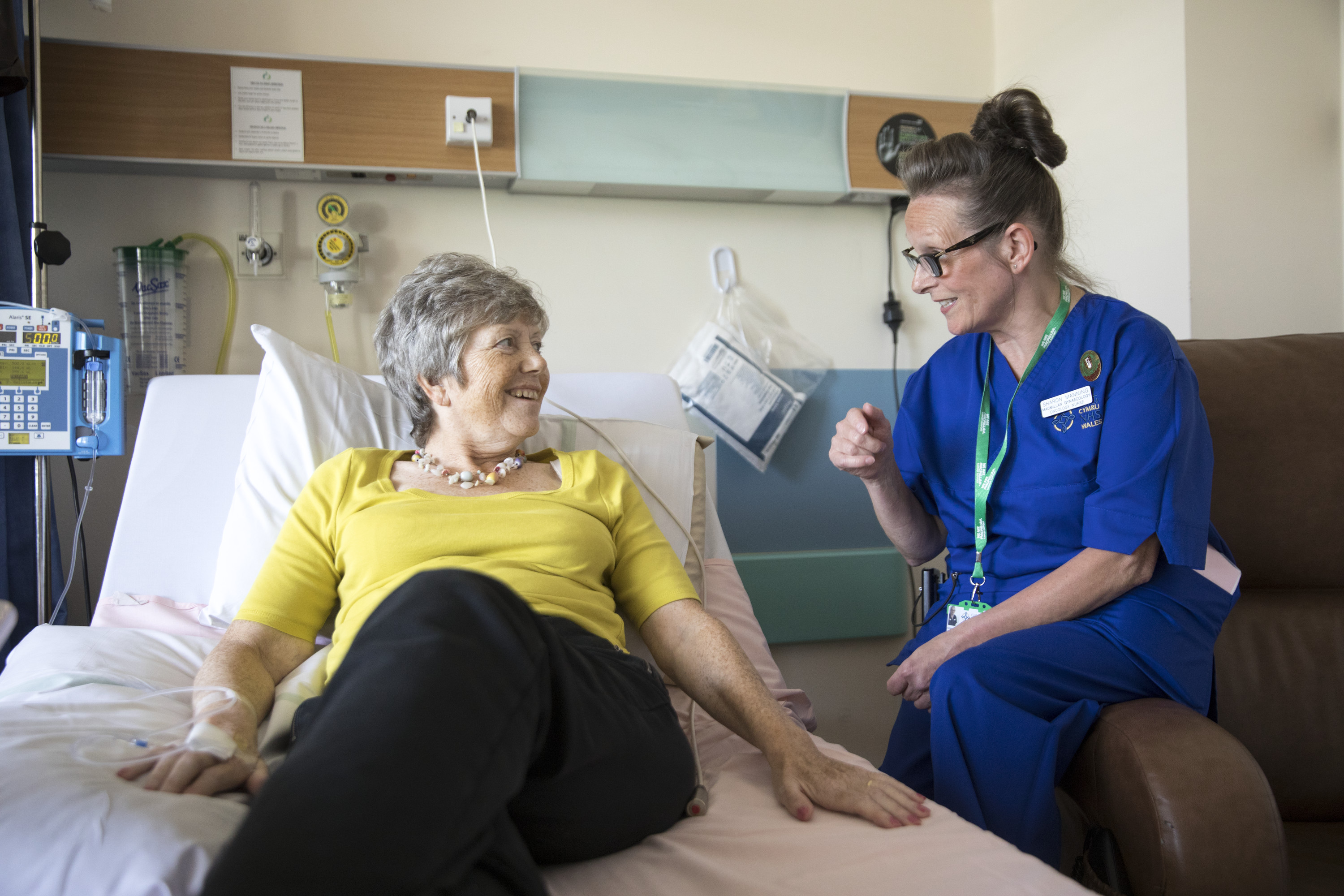 A Macmillan nurse is sitting on the armrest of a chair and speaking to a patient who is laying on a hospital bed. The nurse is wearing a dark blue uniform. The patient is wearing a yellow top and dark trousers. They appear to be in a hospital room. 