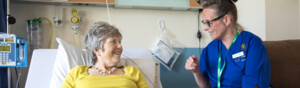 A Macmillan nurse is sitting on the armrest of a chair and speaking to a patient who is laying on a hospital bed. The nurse is wearing a dark blue uniform. The patient is wearing a yellow top and dark trousers. They appear to be in a hospital room. 