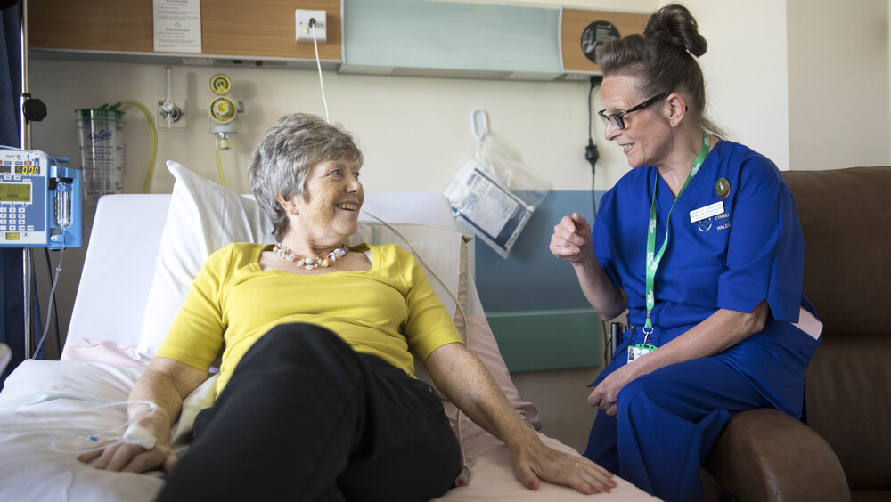 A Macmillan nurse is sitting on the armrest of a chair and speaking to a patient who is laying on a hospital bed. The nurse is wearing a dark blue uniform. The patient is wearing a yellow top and dark trousers. They appear to be in a hospital room. 