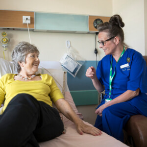 A Macmillan nurse is sitting on the armrest of a chair and speaking to a patient who is laying on a hospital bed. The nurse is wearing a dark blue uniform. The patient is wearing a yellow top and dark trousers. They appear to be in a hospital room. 