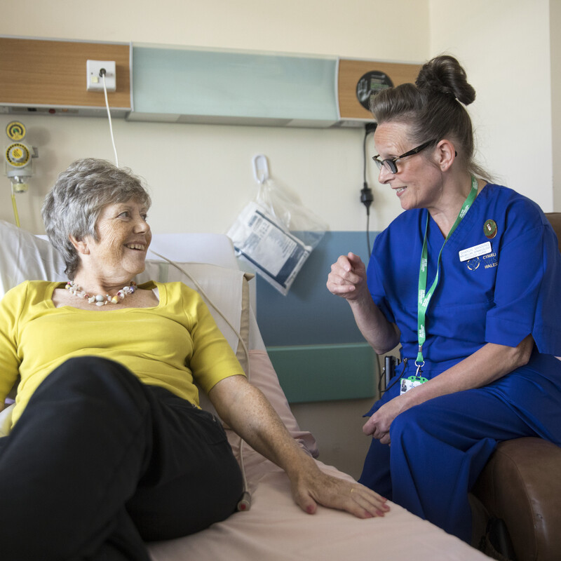 A Macmillan nurse is sitting on the armrest of a chair and speaking to a patient who is laying on a hospital bed. The nurse is wearing a dark blue uniform. The patient is wearing a yellow top and dark trousers. They appear to be in a hospital room. 