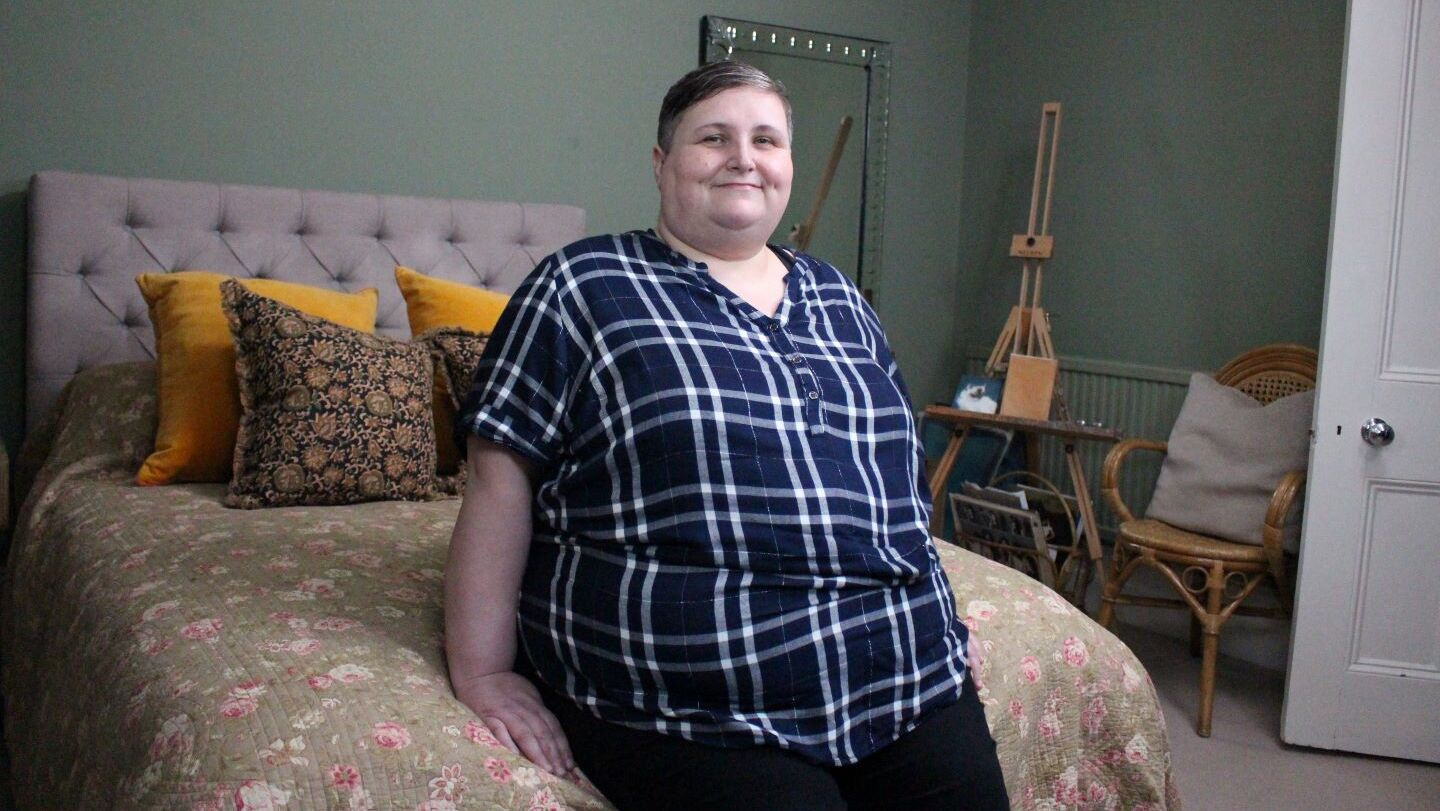 Sarah is sitting on a bed, looking at the camera and smiling.