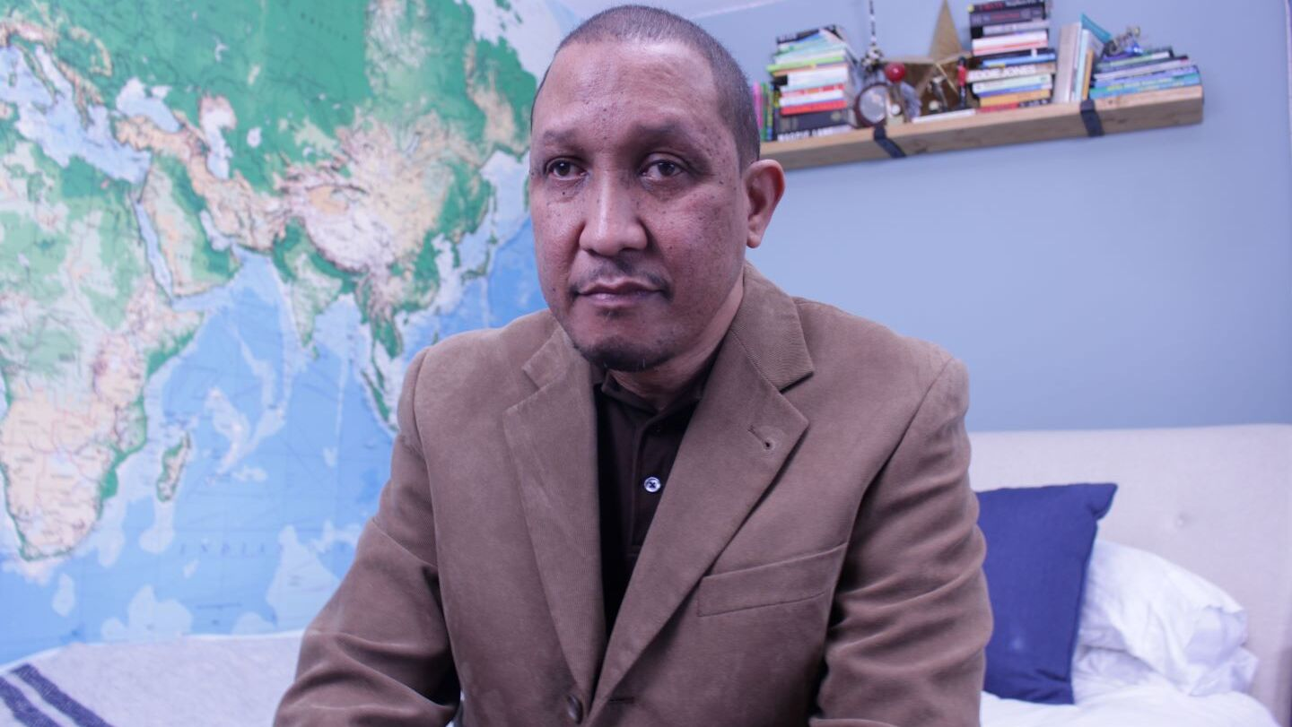 Sean is sitting on a bed and wearing a brown jackert.