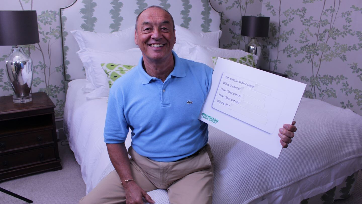 Elvin is sitting on a bed, looking at the camera and smiling. He is holiday a white board with questions about cancer written on it.