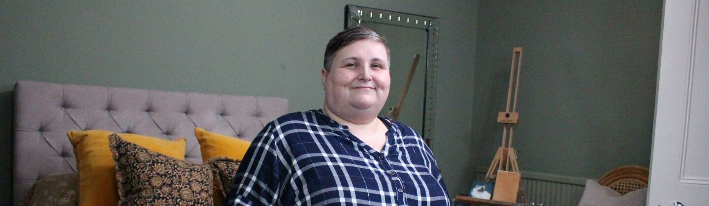 Sarah is sitting on a bed, looking at the camera and smiling.