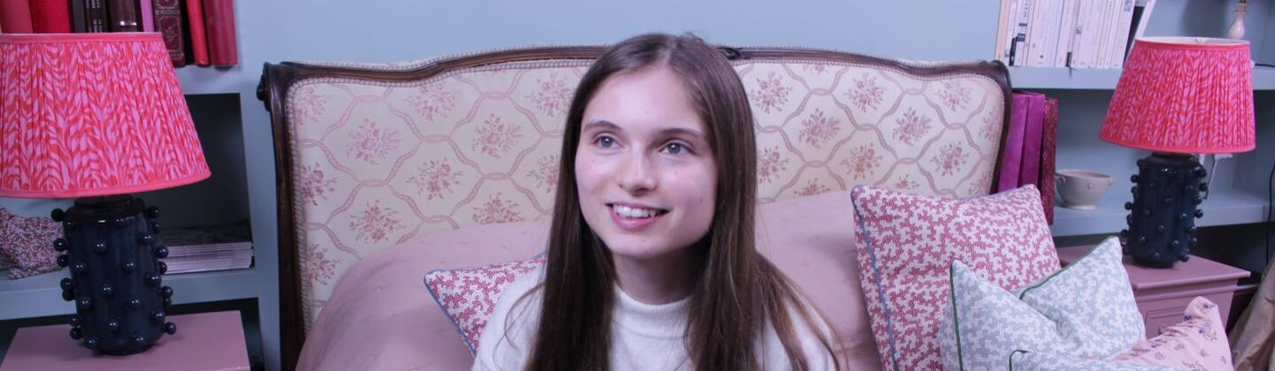 Ellie is sitting on a bed, looking up and smiling.