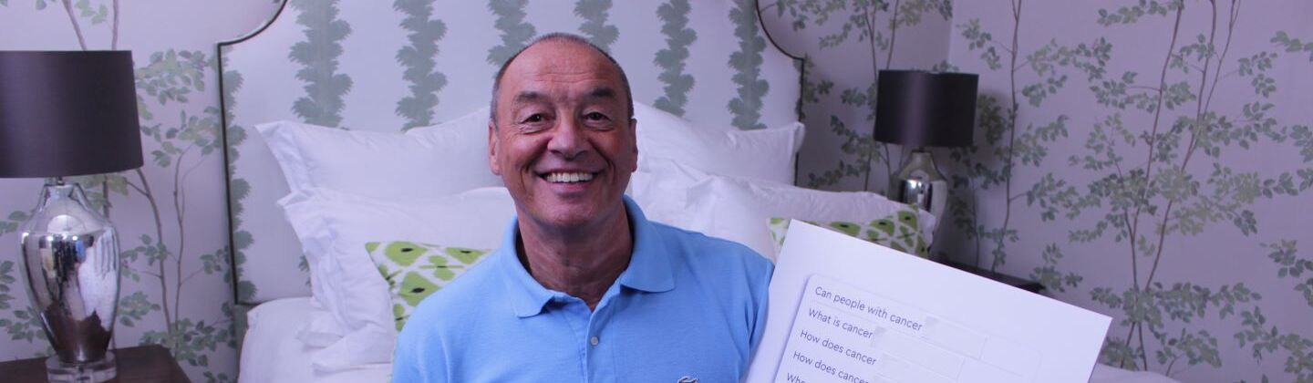 Elvin is sitting on a bed, looking at the camera and smiling. He is holiday a white board with questions about cancer written on it.