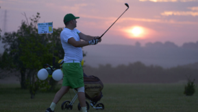A golfer swings his club as the sun sets in the background.