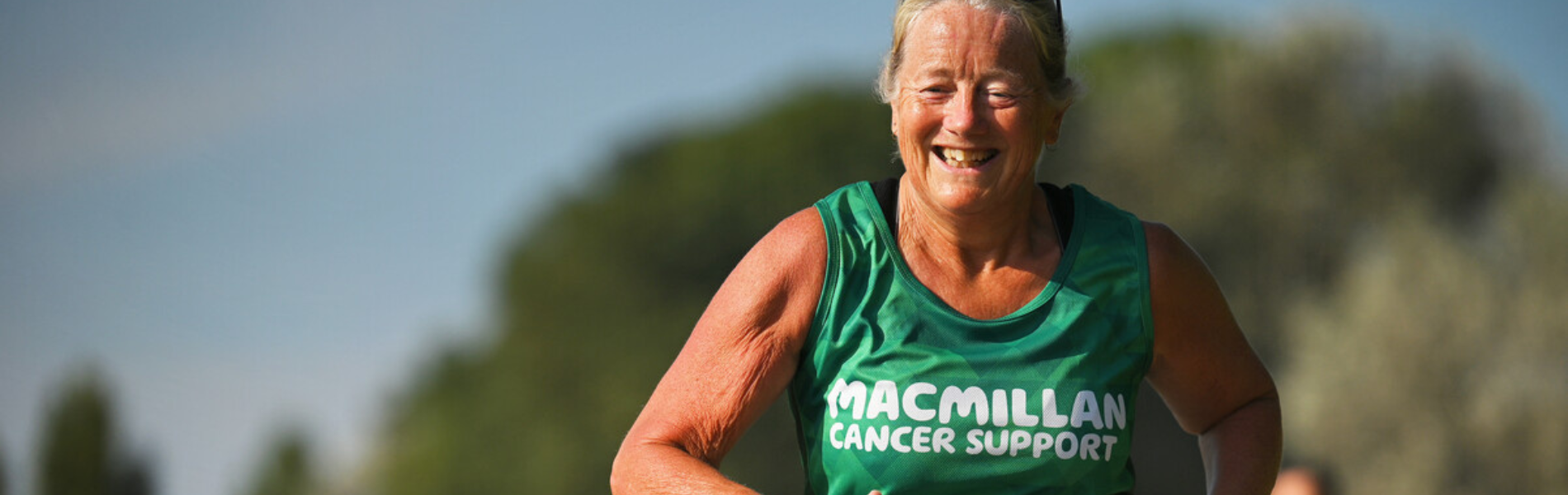 A person wearing a green Macmillan branded vest is running outside. The image is cropped to show them from the elbow up. The background behind them is blurred.