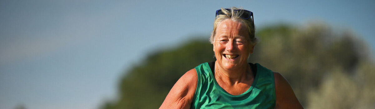 A participant is running outside. They are wearing a green Macmillan branded vest and running shorts. The background behind them is blurry and there appears to be another runner behind the participant.