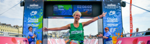 A participant is celebrating after passing the finish line at the Brighton and Hove Triathlon. They are wearing a green Macmillan branded vest, a event number card, and shorts. People are cheering on either side of the participant. 